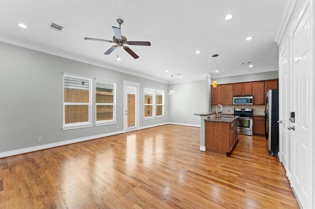 This photo showcases a spacious, open-concept living area with hardwood floors, a ceiling fan, and ample natural light from multiple windows. The modern kitchen features stainless steel appliances, a center island, and wooden cabinetry, creating an inviting space for cooking and entertaining.