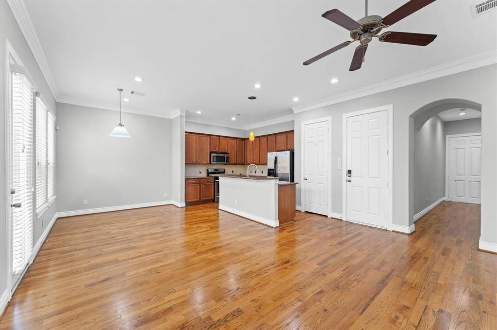 This spacious open-concept living area features hardwood floors, a modern kitchen with stainless steel appliances and an island, ample natural light from large windows, and a ceiling fan for comfort.