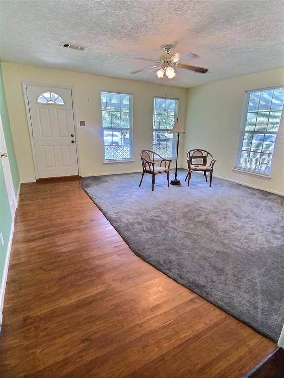 This room features neutral carpeting, two large windows with blinds for natural light, a ceiling fan for air circulation, and a cozy seating nook. Perfect for a bedroom or office space.