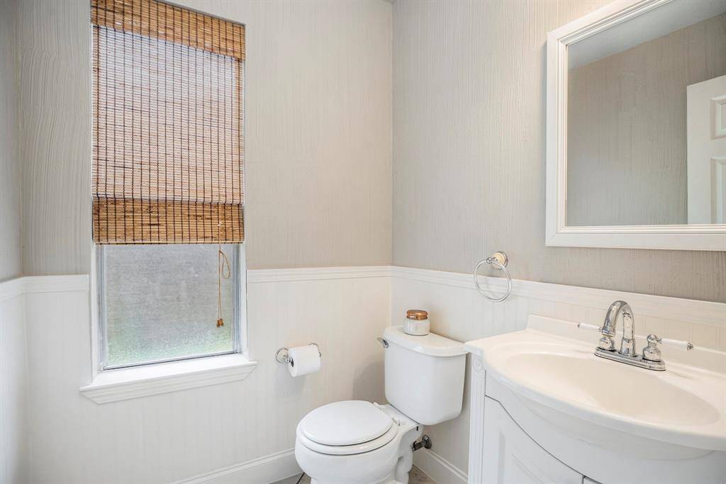The half bath features a furniture-style vanity with a curved front, wainscoting, and soft neutral tones. A window with a woven shade brings in natural light while maintaining privacy.