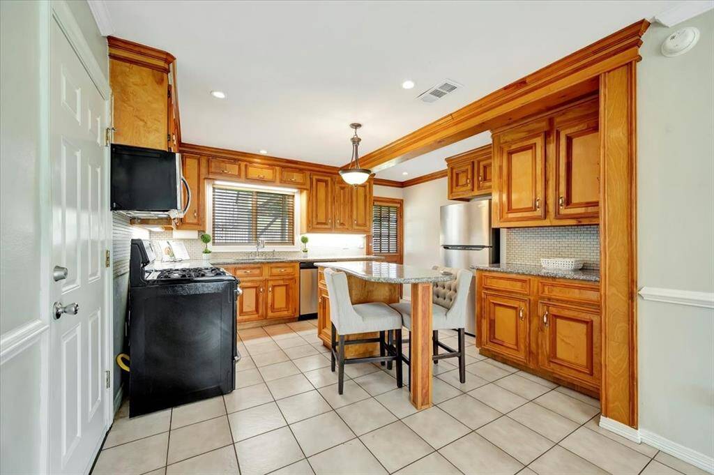 This photo showcases a bright kitchen and dining area with light tile flooring and soft neutral walls. The kitchen features wooden cabinets, stainless steel appliances, and a granite-topped island with seating.