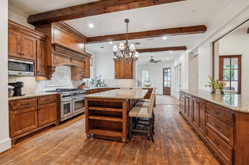This kitchen features warm hardwood flooring, exposed ceiling beams, and custom wood cabinetry throughout. A large central island with granite countertops offers additional workspace and storage. The perimeter counters and backsplash are also finished in complementary tones, and a professional-grade range is set beneath an ornate wood vent hood.