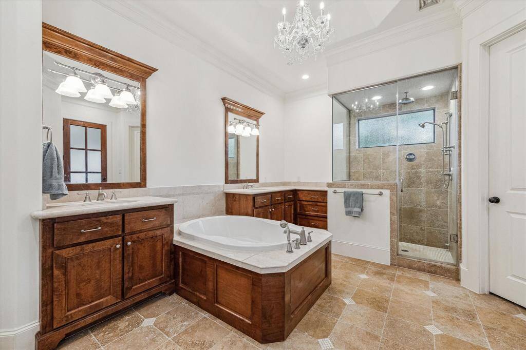This primary bathroom features a spacious layout with separate vanities, a large walk-in shower with dual shower heads, and a centrally located soaking tub. The room is well-lit with both natural and recessed lighting, and it includes a high ceiling that enhances the open feel. Ample counter space and storage are built into the design for functionality.