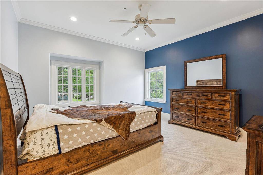 This secondary bedroom features high ceilings, crown molding, and large windows that provide natural light and views of the outdoors. The space is carpeted and includes recessed lighting and a ceiling fan for comfort.