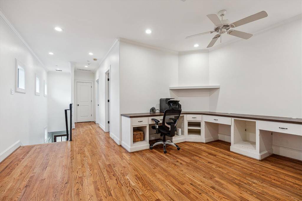 This upstairs work area features built-in desk space with ample countertop surface and multiple storage compartments. It has recessed lighting, hardwood floors, and a ceiling fan, with natural light coming from a series of small windows along the hallway. The layout is open and functional, ideal for a study or home office setup.