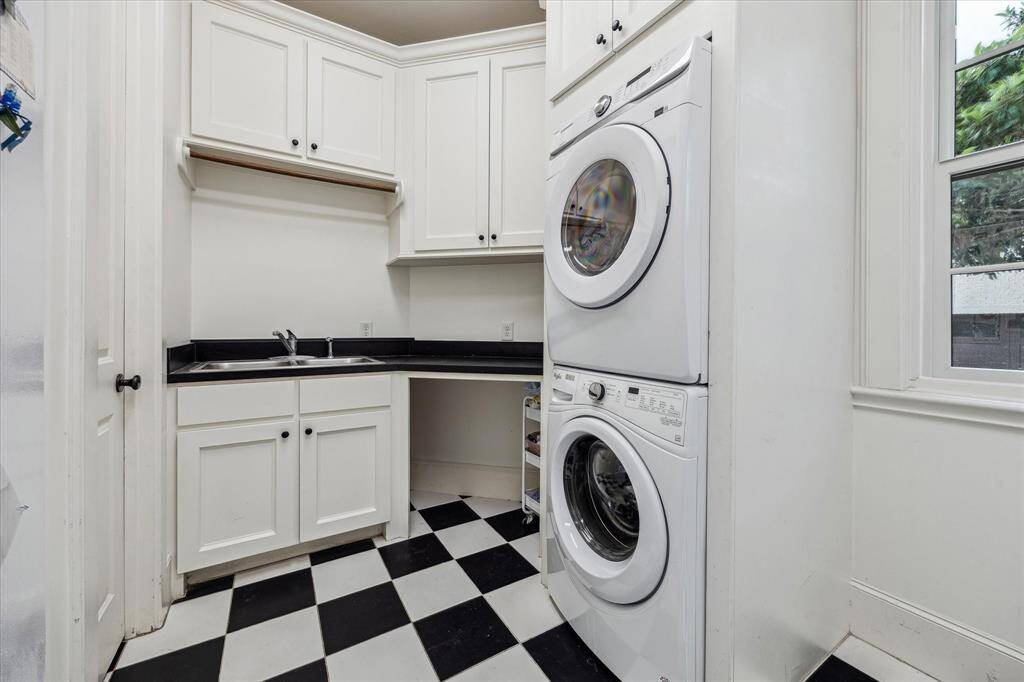 This utility room includes a stacked washer and dryer, a deep sink with counter space, upper and lower cabinetry for storage, and a hanging rod for air-drying clothes. A window provides natural light, and the space is finished with a checkered tile floor.