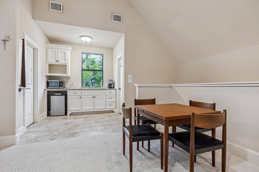 This garage apartment includes a compact kitchenette with upper and lower cabinetry, a sink, and a window that provides natural light. The space features a combination of tiled and carpeted flooring, central air vents, and a sloped ceiling that adds architectural interest. A nearby door likely leads to a private bathroom or storage area.