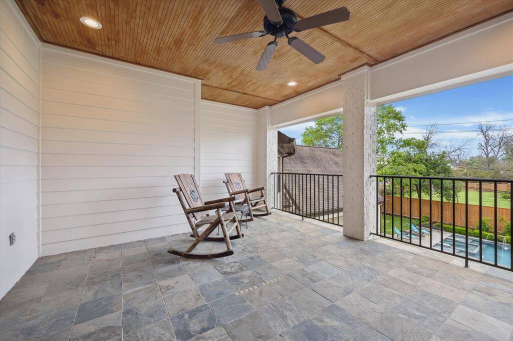 This second-floor covered balcony features a tiled floor, wood-paneled ceiling with recessed lighting, and a ceiling fan for airflow. It offers a spacious layout and open views of the backyard, including the pool area and surrounding greenery, with a protective railing along the edge.