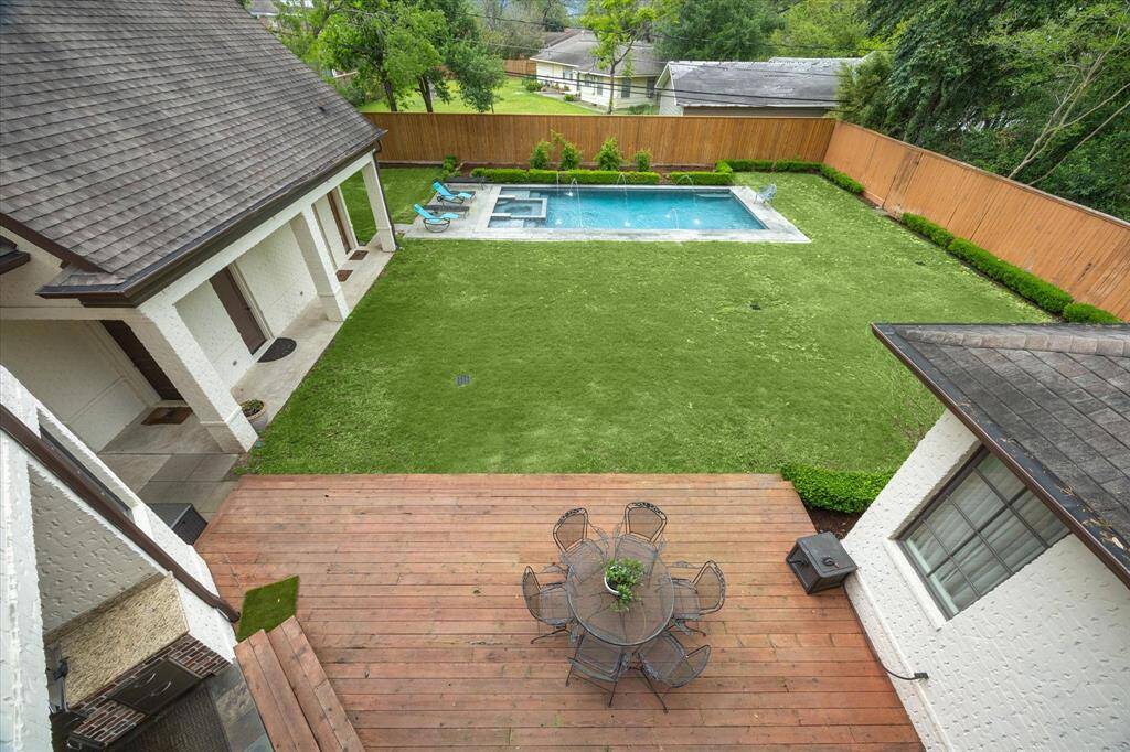 This view of the backyard shows a large grassy lawn bordered by fencing and neatly trimmed hedges. A swimming pool with water features and an attached spa is located toward the back of the yard. A spacious wooden deck sits adjacent to the house, providing access to both the yard and pool area.