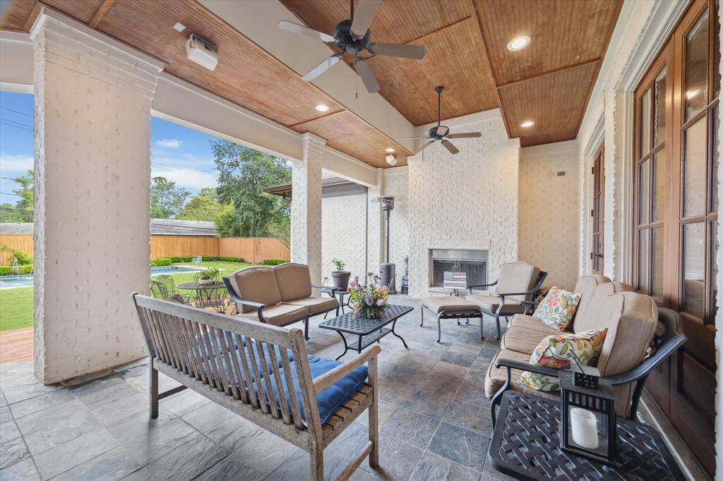 This first-floor covered patio features a tiled floor, wood-paneled ceiling with recessed lighting, and ceiling fans for ventilation. It includes a built-in fireplace and is open to the backyard, offering a sheltered space for outdoor relaxation and entertaining.