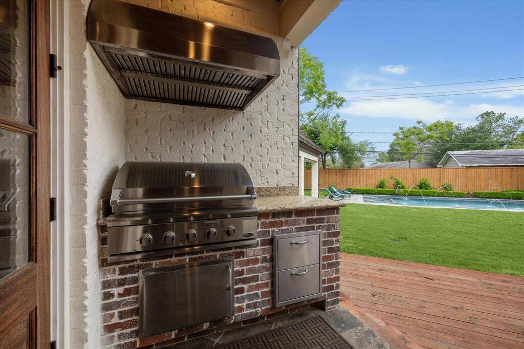 This outdoor kitchen features a built-in stainless steel grill with a vent hood, granite countertops, and brick base construction. It includes storage drawers and cabinetry, and is positioned near the backyard deck with views of the pool and lawn area.
