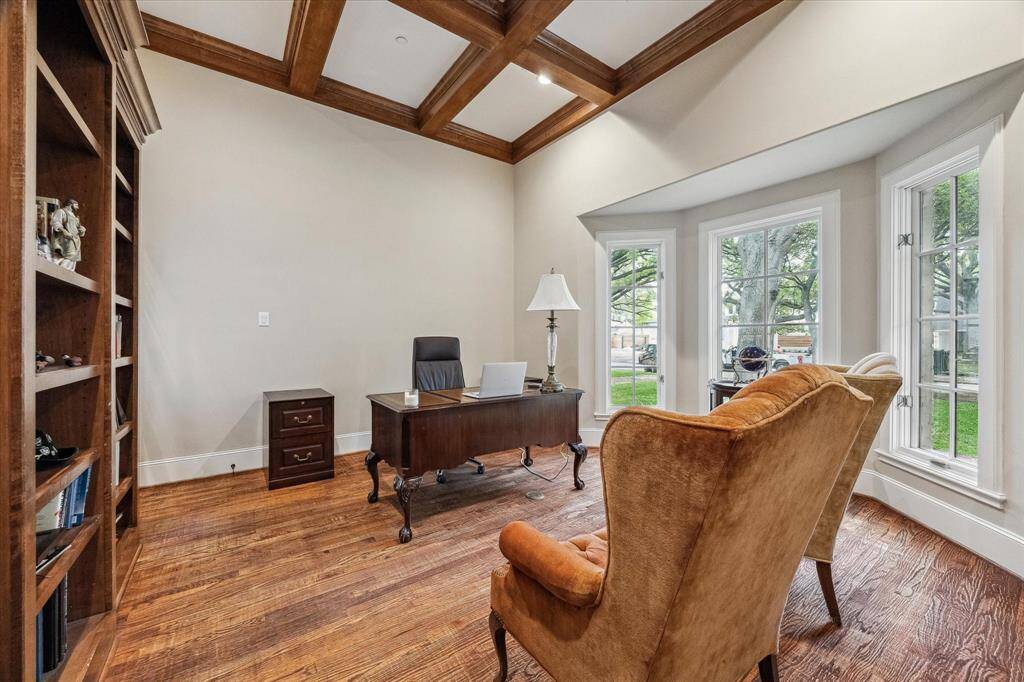 This formal study features rich hardwood floors, a vaulted ceiling with an elegant wood-beamed coffered design, and neutral-toned walls that enhance the natural light. A large bay window with multiple panes offers a scenic view of the front yard, while built-in shelving provides architectural character and functionality. The space is both bright and refined, ideal for a home office or library.