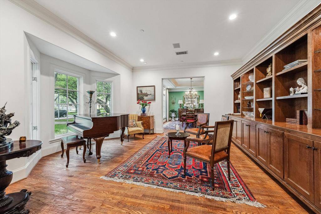 This formal living room features wide-plank hardwood flooring, crisp white walls, and crown molding for a refined finish. Large bay windows flood the space with natural light and offer views of the front lawn. Recessed lighting and an open layout enhance the airy feel, while custom built-in shelving along one wall adds both architectural detail and functional storage. The room opens directly into a formal dining area, creating a seamless flow for entertaining.