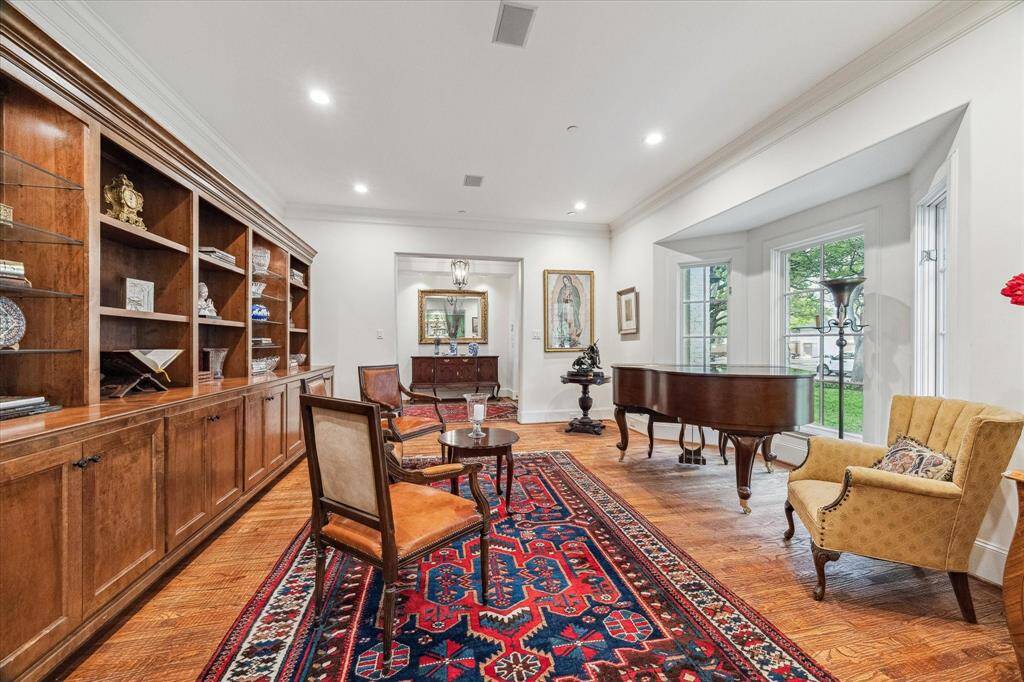 This formal living room is defined by its warm hardwood flooring, tall ceilings with recessed lighting, and a wall of custom built-in cabinetry offering ample shelving and storage. A large bay window brings in abundant natural light and provides views of the front yard. The room connects directly to the foyer, creating a welcoming and open flow through the home.