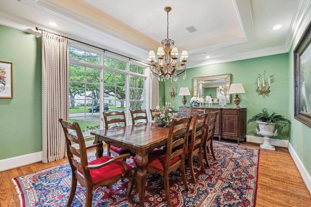 This formal dining room features rich hardwood flooring, a large picture window that fills the space with natural light, and elegant crown molding throughout. The ceiling is recessed with a tray design, adding depth and architectural interest. Soft recessed lighting enhances the ambiance, making it a bright and inviting setting for gatherings.