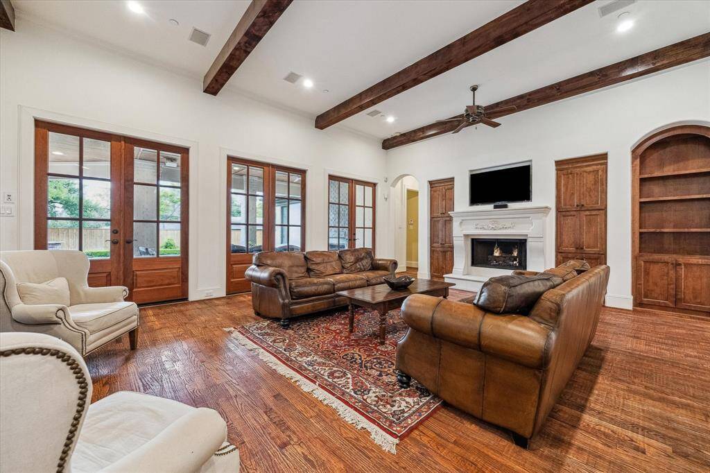 This spacious family room features warm hardwood flooring, vaulted ceilings with exposed wooden beams, and abundant natural light from a wall of glass-paneled French doors leading to the outdoor living area. A central fireplace with a detailed mantel anchors the room, flanked by custom built-in cabinetry and shelving for storage and display.