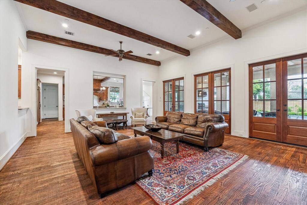 This inviting family room showcases warm hardwood flooring, white walls, and exposed wooden ceiling beams that add rustic charm. Multiple sets of French doors line one wall, offering abundant natural light and access to the outdoor living space. The room connects seamlessly to the kitchen through a large open pass-through and adjacent doorway, enhancing flow and functionality for everyday living and entertaining.
