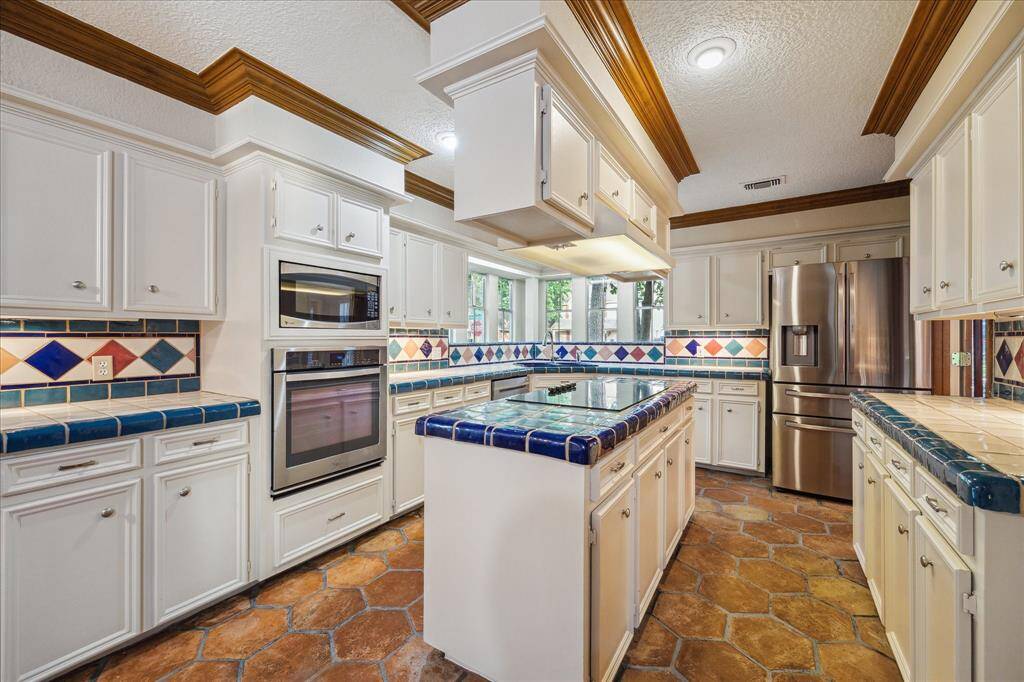 This kitchen features a spacious layout with white cabinetry, colorful tile countertops, and a central island with glass cooktop.  Modern appliances, ample storage, and a stylish tiled backsplash, creates a bright and inviting space for cooking and entertaining.