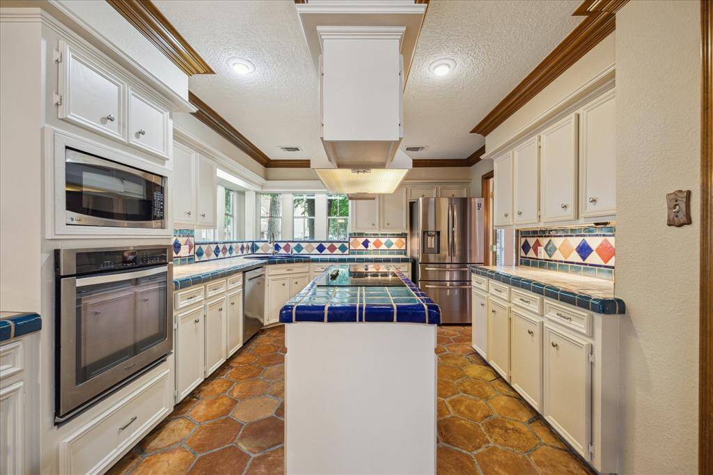 This kitchen features classic white cabinetry, vibrant tile countertops, and a multi-colored tile backsplash. It includes modern stainless steel appliances, a central island, and saltillo tile flooring, creating a warm and inviting cooking space.