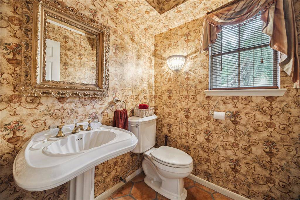 Elegant vintage wallpaper and a classic pedestal sink with brass fixtures. A decorative mirror and soft window drapes add charm, while the salitto floor complements the warm tones.