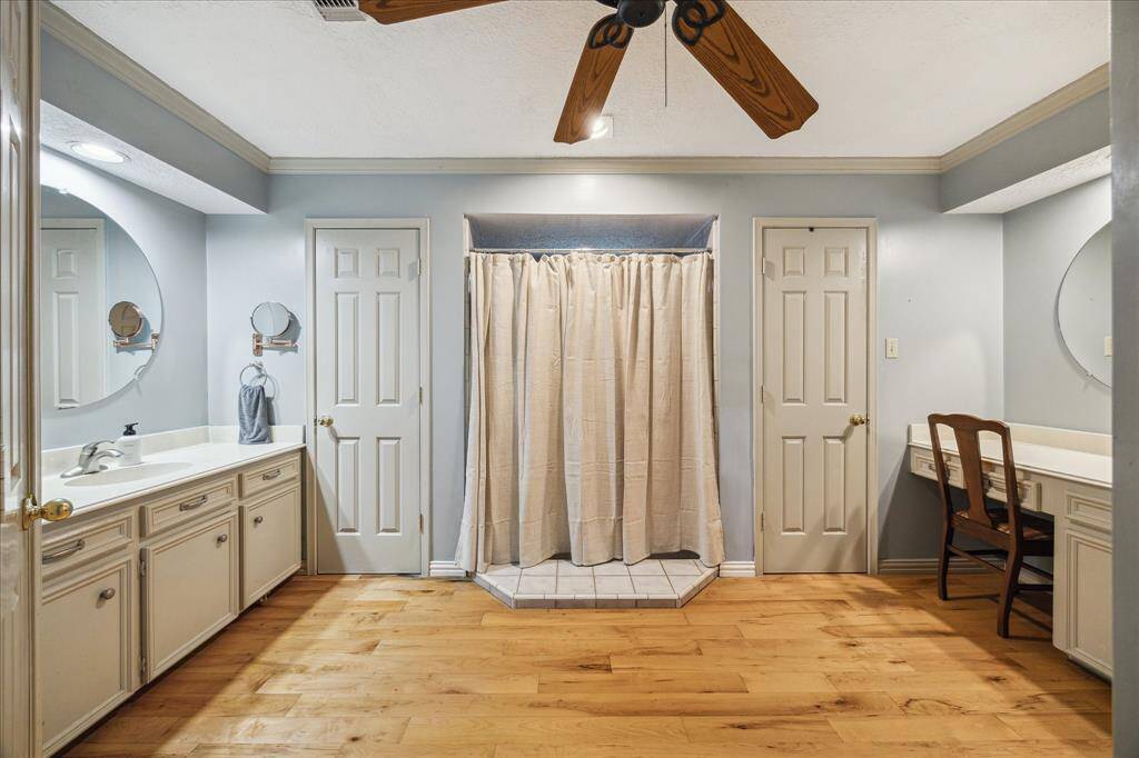 Primary ensuite bathroom features dual sinks with large mirrors, ample cabinet storage, and a central shower area. It has hardwood flooring, a ceiling fan, and a vanity area with a chair, offering a blend of functionality and comfort.