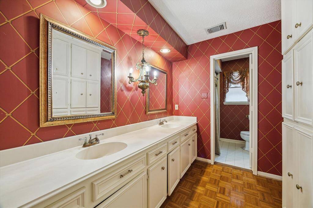 This bathroom features a long vanity with dual sinks and classic white cabinetry, dual sinks and tub/shower combo. It has rich, red diamond-patterned wallpaper and elegant lighting fixtures, adding a touch of luxury.