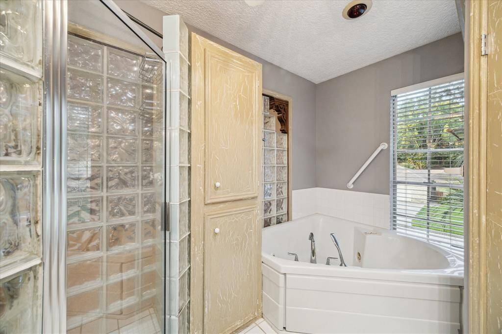 Bathroom features a glass block shower, a corner jetted tub, and a large window offering natural light. The space includes textured cabinets for storage and a neutral color scheme.