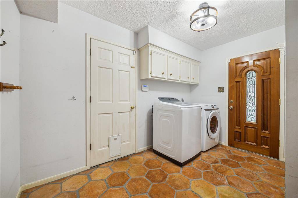 First floor laundry room with overhead cabinets for storage, and a charming wooden door with decorative glass. The space has saltillo tile flooring and a modern ceiling light fixture.  Two doors, one going outside and the other going into the garage.