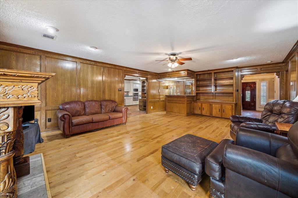 Spacious living room with warm wood paneling, built-in shelving, and a cozy fireplace. Features hardwood flooring, ceiling fan, and ample natural light. Adjacent to the kitchen, ideal for entertaining.