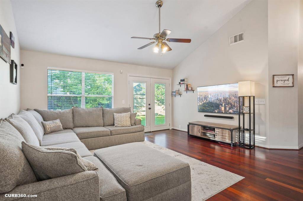 Spacious and inviting living room featuring vaulted ceilings and rich wooden floors. Abundant natural light streams through large windows, highlighting a cozy sectional perfect for relaxation. Enjoy seamless indoor-outdoor flow with direct access to the lush backyard.