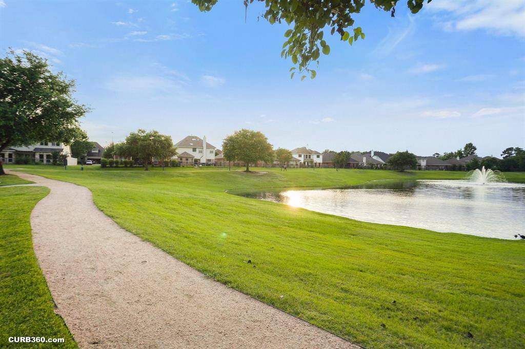 Discover serene living with this picturesque view: a lush green walkway alongside a tranquil pond, complete with a charming fountain. Perfect for peaceful strolls and enjoying nature's beauty.