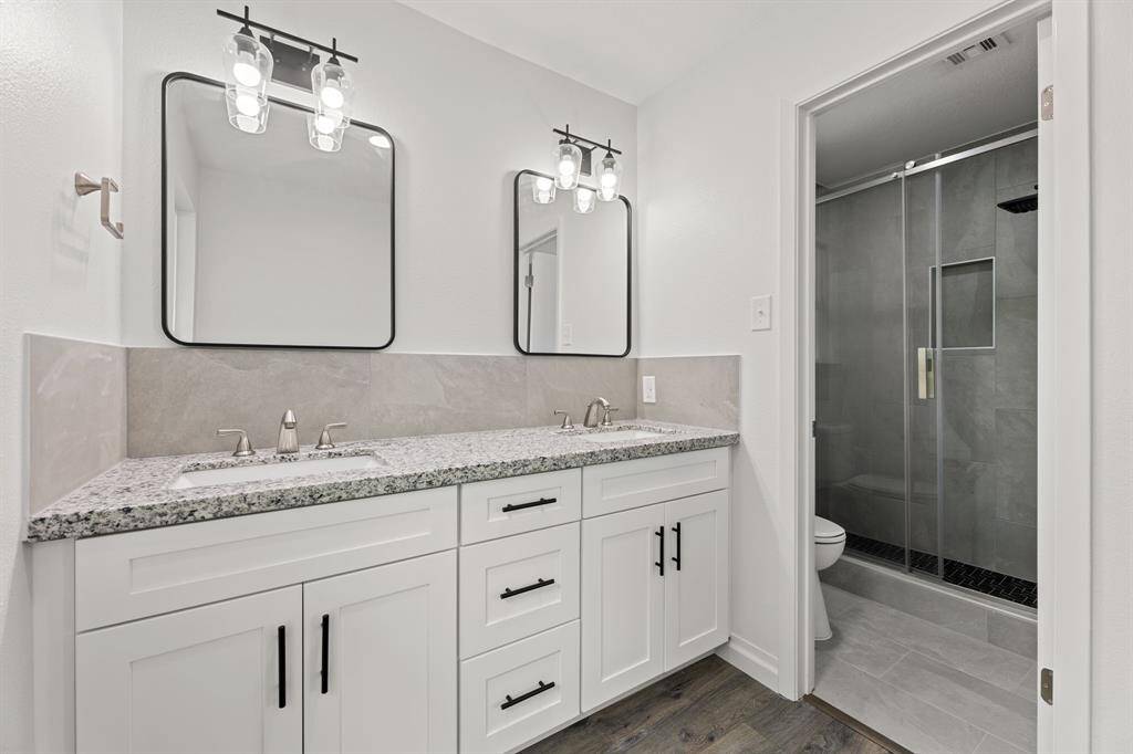 This modern bathroom features a double vanity with granite countertops, sleek black fixtures, and elegant lighting. It includes a spacious walk-in shower with glass doors and contemporary tile work.
