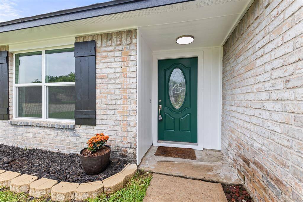Charming entry with a green front door and oval glass insert, framed by light brick siding. Features a cozy covered porch area, a large window with black shutters, and a tidy garden bed with potted flowers.