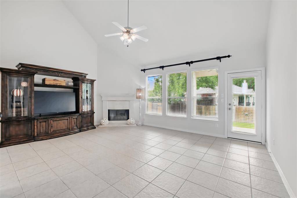 Spacious and bright living room featuring soaring ceilings, a wall of windows for abundant natural light, and a cozy corner fireplace with a classic white mantle. The neutral tile flooring and fresh paint create a perfect blank slate, ready to complement any style. French door leads to the backyard for seamless indoor-outdoor living.