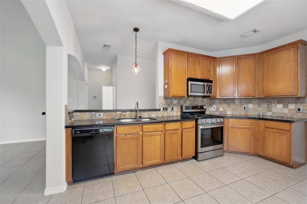 This spacious kitchen features warm wood cabinetry, granite countertops, and a stylish stone tile backsplash that adds texture and character. Stainless steel appliances, including a gas range and built-in microwave, offer modern convenience, while a pendant light over the sink brings a touch of charm. The open layout connects effortlessly to adjacent living spaces, making it ideal for everyday living and entertaining.