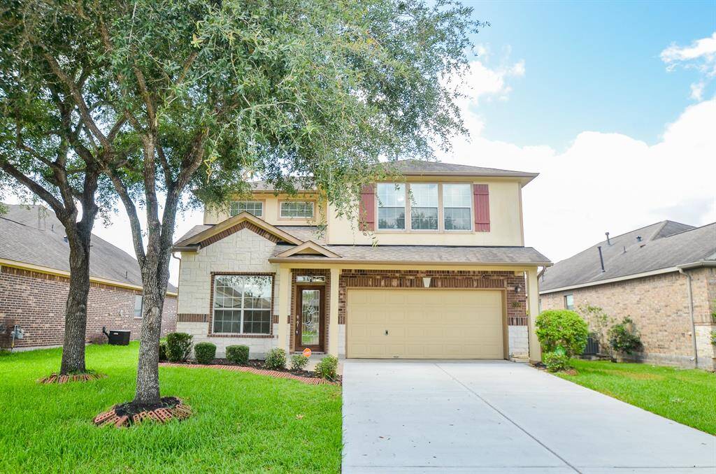 This is a two-story suburban home with a beige exterior and shutters, featuring a two-car garage. It has a well-maintained front lawn with mature trees and a paved driveway. The house is set in a neighborhood with similar brick homes.