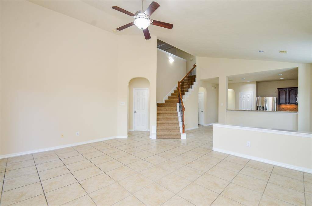 This spacious living area features high ceilings, neutral tile flooring, and a ceiling fan. An open staircase leads to the upper level, and the room flows into a modern kitchen with stainless steel appliances and dark wood cabinetry. Perfect for comfortable family living.