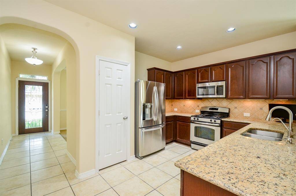 This kitchen features granite countertops, stainless steel appliances, and rich wood cabinetry. It has a tiled floor and an open layout, leading to a well-lit entrance hallway with a modern front door.