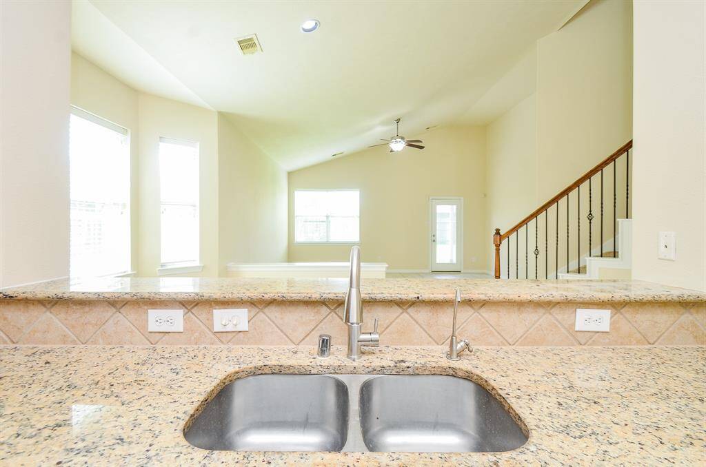 Bright and open living area with high ceilings, visible from the kitchen featuring granite countertops and a double sink. Stairs with decorative railing lead to the upper level, and large windows provide ample natural light.
