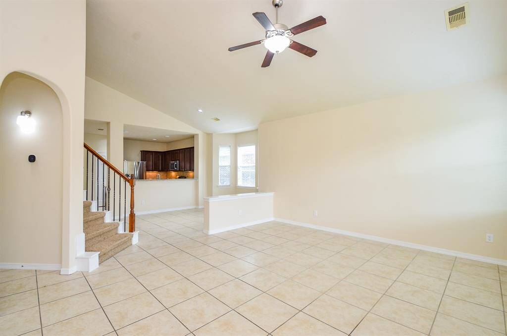 Spacious living area with tiled flooring, high ceiling, and ceiling fan. Features an open layout leading to a modern kitchen with stainless steel appliances. Carpeted stairs add warmth and lead to the upper level. Abundant natural light from large windows.