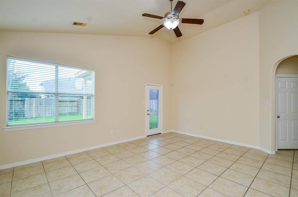 This spacious living room features high ceilings with a ceiling fan, large windows for natural light, tile flooring, and neutral walls. It has a door leading to the backyard, creating a bright and airy living space.