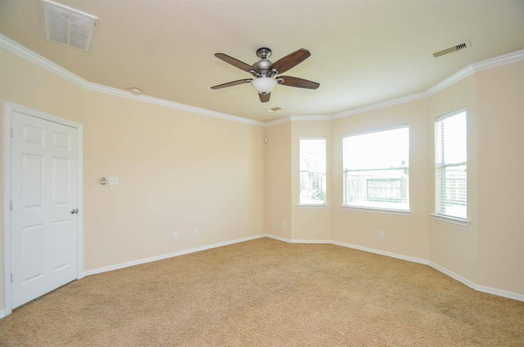 This bright primary bedroom features neutral walls, carpeted flooring, a ceiling fan, and ample natural light from three windows. Ideal for a cozy living space or bedroom.