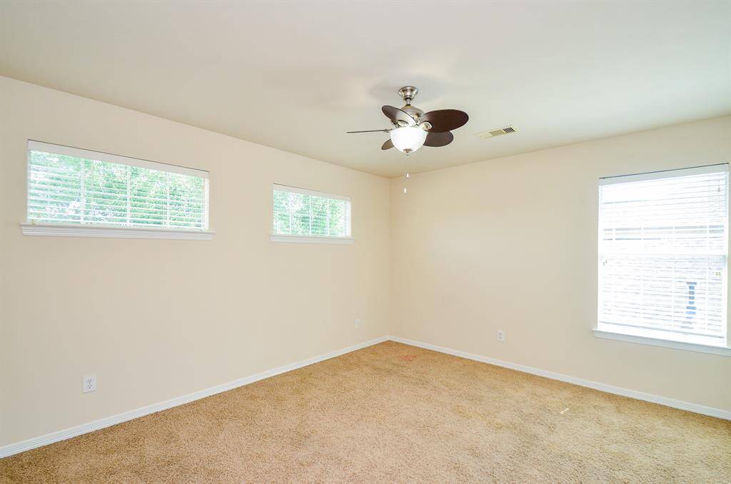This secondary bedroom is spacious with carpeted flooring, neutral walls, and ample natural light from multiple windows. It features a ceiling fan and overhead lighting, making it bright and airy.