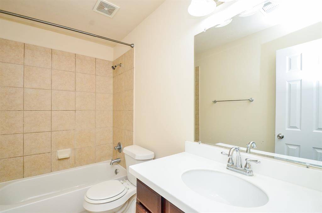 This secondary bathroom features a clean, modern design with beige tiled walls surrounding a bathtub, a white vanity with a sleek faucet, and a large mirror. The space is well-lit and includes a towel rack and standard fixtures.