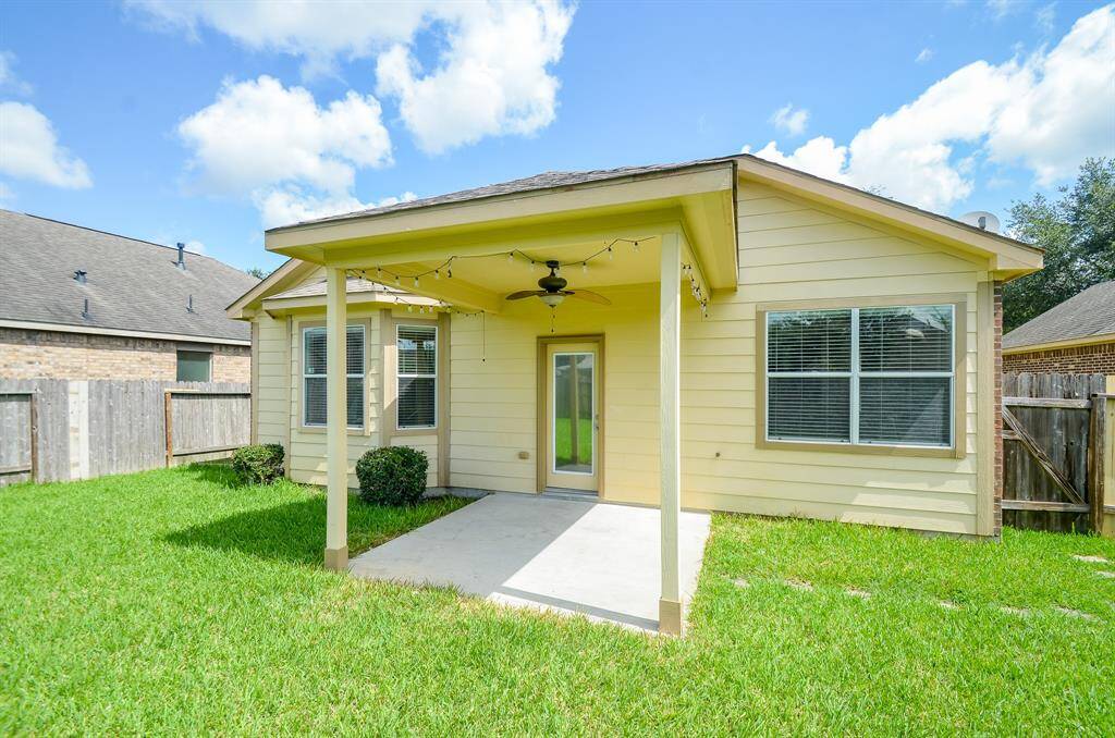 This photo shows a backyard with a small covered patio attached to the house. The patio has a ceiling fan and string lights, overlooking a grassy lawn enclosed by a wooden fence. The house features light-color siding and multiple windows, offering a cozy outdoor space.