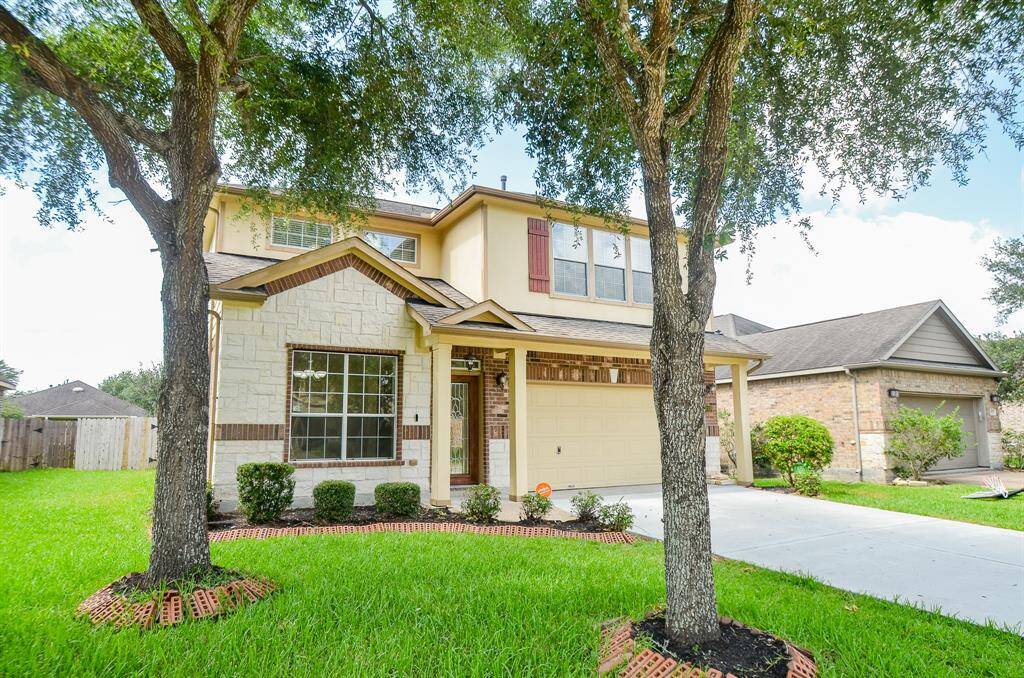 This charming two-story home features a stone and brick facade with a welcoming front porch. It is framed by mature trees and has a well-maintained lawn with a paved driveway leading to the garage. Perfect for a family seeking a suburban setting.