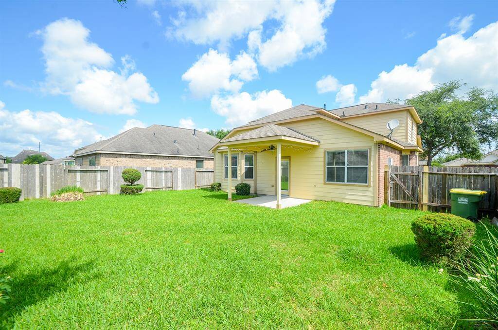 This photo showcases a charming backyard with a spacious, well-maintained lawn, bordered by a wooden fence. The house features beige siding with a covered patio, perfect for outdoor relaxation. The surrounding landscape includes neatly trimmed shrubs and clear blue skies, creating a serene atmosphere.