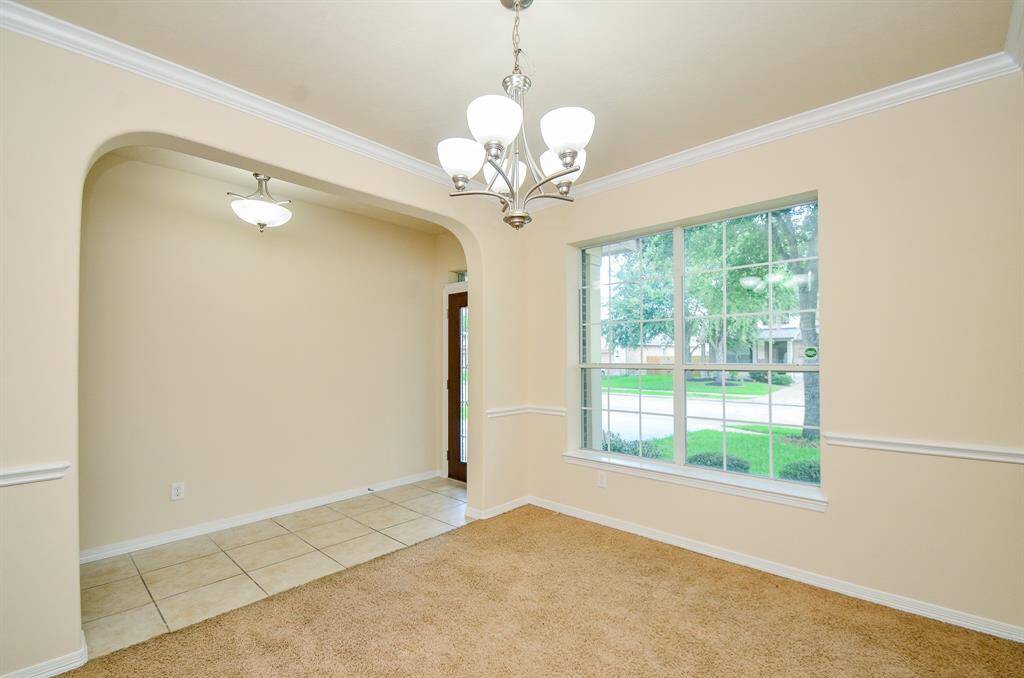 A bright dining area with large windows offering plenty of natural light. It features a modern chandelier, neutral-colored walls, and a mix of carpet and tile flooring. The space is open and inviting, ideal for gatherings.
