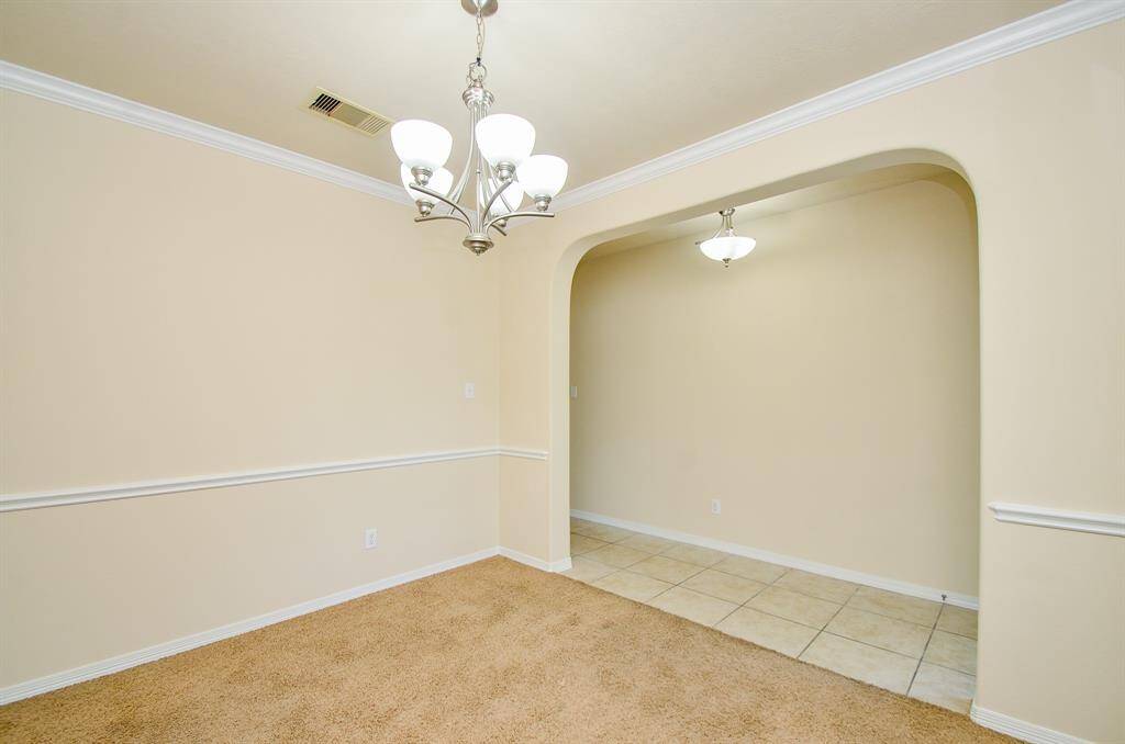 This photo shows a bright dining area with soft beige walls, a plush carpet, and a stylish chandelier. An adjacent space with tiled flooring features a matching light fixture, separated by an arched entryway, enhancing the elegant open layout.