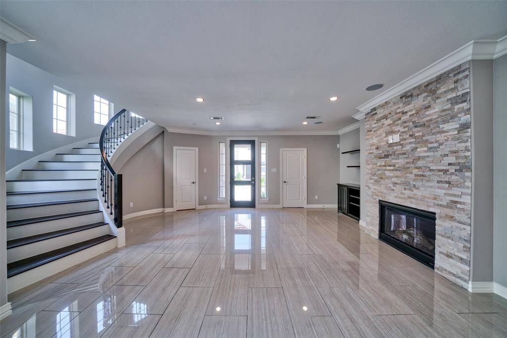 The spacious living room features tile floors, a stone fireplace, and a curved staircase. Large windows provide ample natural light, and the neutral color palette enhances the airy feel.
